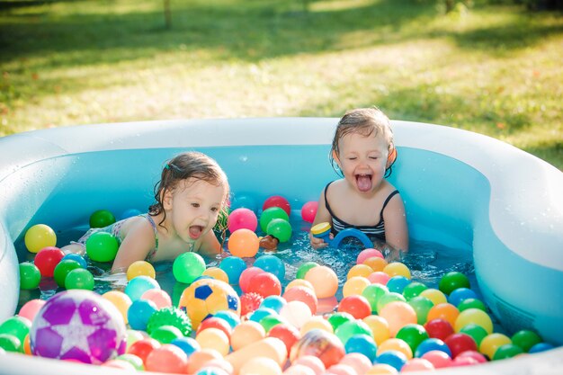 As duas meninas de dois anos de idade brincando com brinquedos na piscina inflável no dia ensolarado de verão