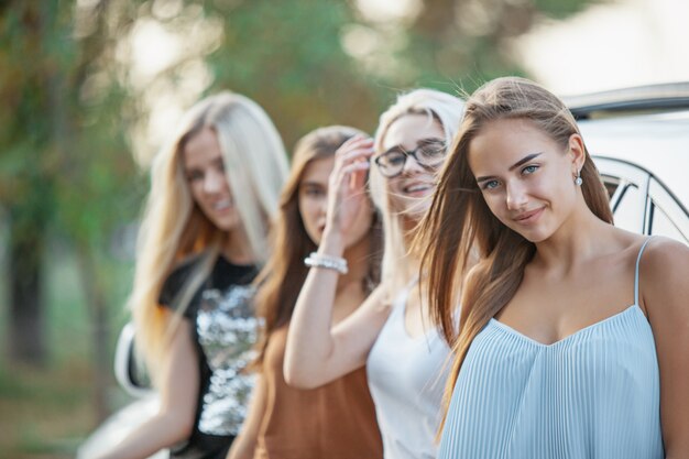 As jovens mulheres de pé e sorrindo perto do carro ao ar livre. O estilo de vida, viagens, aventura e conceito de amizade feminina