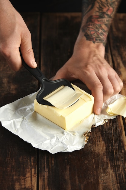 Foto grátis as mãos do homem tatuado usam uma faca especial para picar uma fatia bem fina de manteiga, tudo em uma mesa de madeira rústica