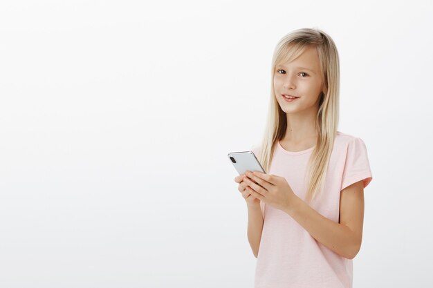 As meninas podem usar gadgets melhor do que os pais. Retrato de criança adorável e confiante com lindos olhos em uma camiseta rosa, segurando um smartphone e sorrindo