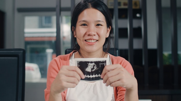 As mulheres gravidas asiáticas novas mostram e olhando o bebê da foto do ultrassom na barriga. mãe, sentindo-se feliz sorrindo pacífica enquanto cuida criança sentada na mesa na sala de estar em casa de manhã.