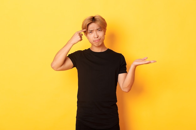 Foto grátis asiático decepcionado com cabelo loiro, levantando a mão intrigado e repreendendo alguém que agia de estúpido, parado na parede amarela