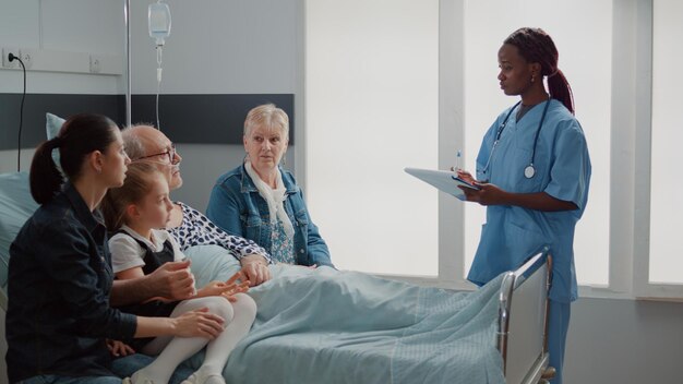 Assistente afro-americano explicando medicação para um homem idoso e visitantes na enfermaria do hospital. Especialista em conversa com a família do paciente doente sobre tratamento e cuidados de saúde. Enfermeira dando conselhos