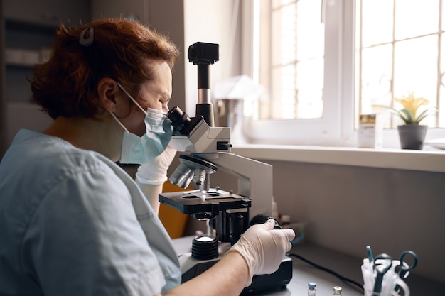 Assistente de laboratório maduro na máscara olha para o microscópio pesquisando amostra no local de trabalho