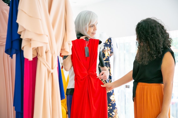 Assistente de loja mostrando vestido de festa com etiqueta para o cliente entre prateleiras com roupas. Mulher escolhendo roupas de noite. Vista lateral. Loja de moda ou conceito de varejo