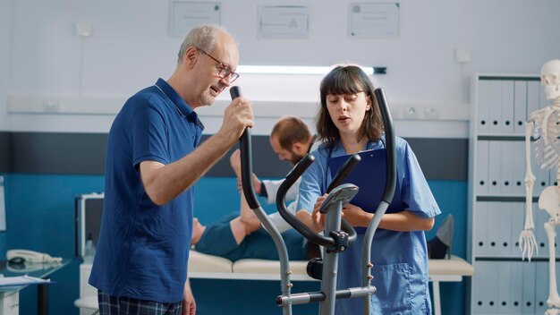 Assistente médico apoiando o homem sênior fazendo fisioterapia, exercitando-se em bicicleta estacionária. Enfermeira ajudando a pessoa idosa a frequentar fisioterapia de reabilitação no gabinete.