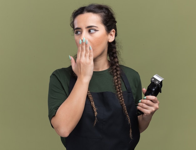 Foto grátis assustada ao olhar para o lado da jovem barbeira de uniforme segurando a tesoura de cabelo coberta com a boca isolada na parede verde oliva