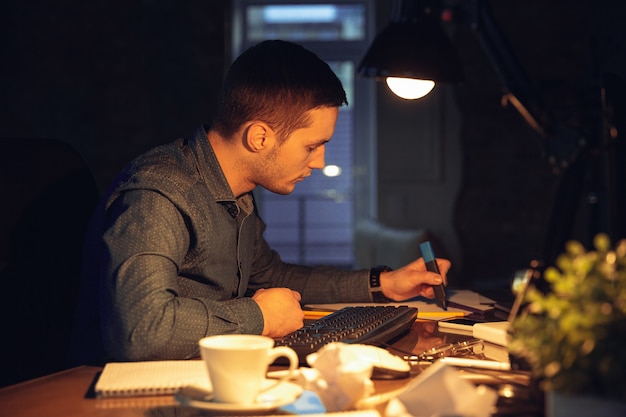 Atendido. Homem trabalhando sozinho no escritório durante a quarentena do coronavírus ou COVID-19, permanecendo até tarde da noite. Jovem empresário, gerente fazendo tarefas com smartphone, laptop, tablet no espaço de trabalho vazio.