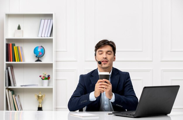 Atendimento ao cliente bonitão com fone de ouvido e laptop de terno fechando os olhos e segurando um café