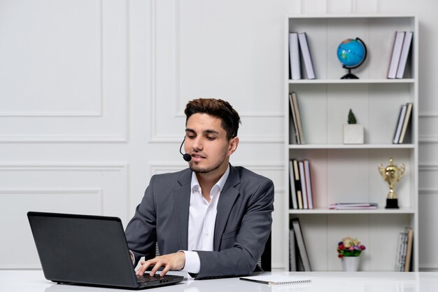 Atendimento ao cliente bonitão inteligente com laptop em blazer de escritório cinza digitando um pedido