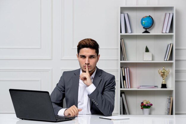 Atendimento ao cliente bonitão inteligente com laptop em blazer de escritório cinza mostrando gesto de silêncio