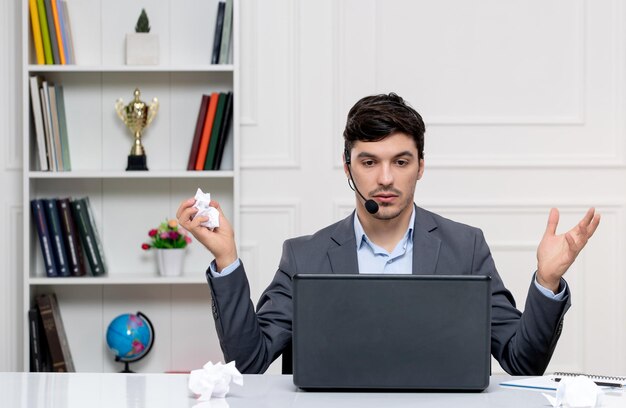 Atendimento ao cliente bonitinho de terno cinza com computador e fone de ouvido acenando com as mãos e segurando um papel