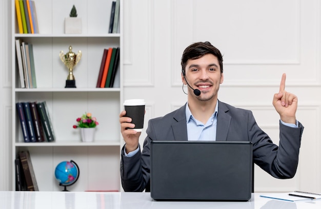 Foto grátis atendimento ao cliente bonitinho de terno cinza com computador e fone de ouvido apontando para cima e sorrindo