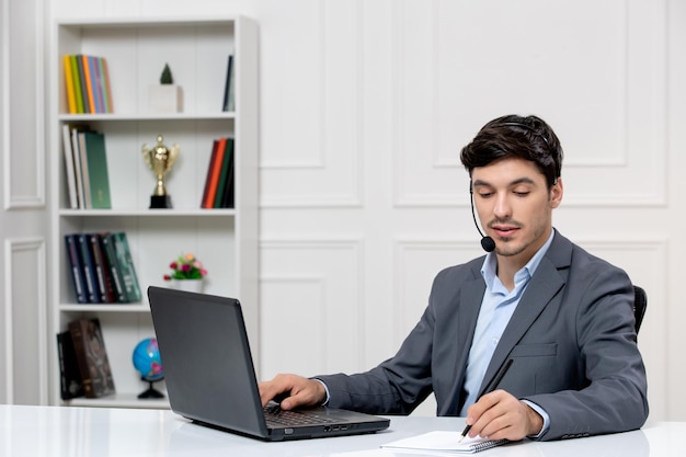 Atendimento ao cliente bonitinho de terno cinza com computador e fone de ouvido escrevendo notas