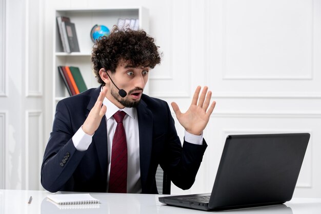 Atendimento ao cliente jovem bonito em terno de escritório com laptop e fone de ouvido chocado acenando as mãos