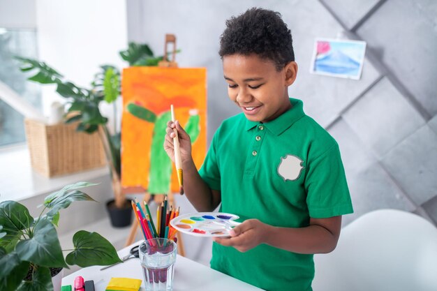 Atividade interessante. Menino de pele escura sorridente em idade escolar primária em camiseta verde, olhando segurando o pincel sobre a paleta com tintas em pé perto da mesa com lápis e copo de água