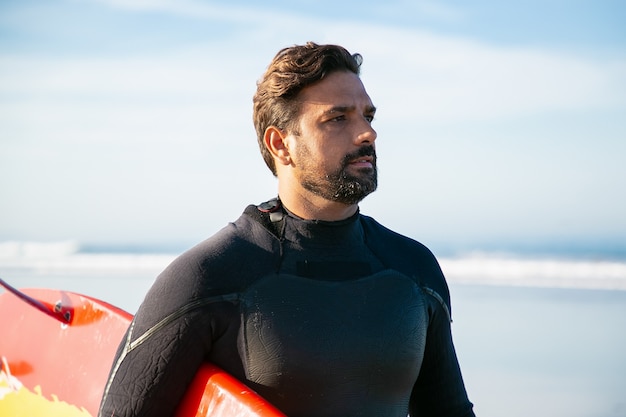 Atleta caucasiana com roupa de neoprene segurando a prancha de surf e olhando para longe