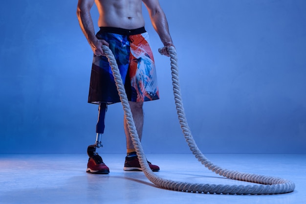 Atleta com deficiência ou amputado isolado na parede azul. Esportista profissional masculino com treinamento de prótese de perna com cordas em neon. Esporte para deficientes físicos e superação, conceito de bem-estar.