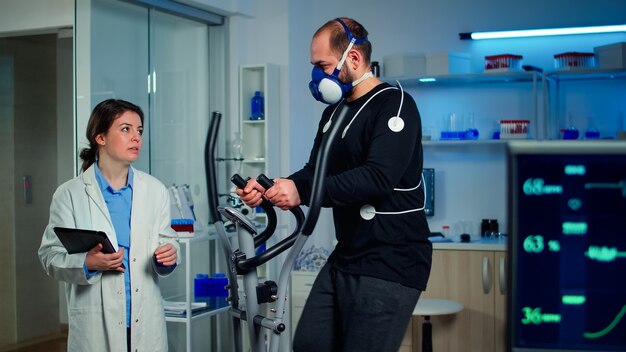 Atleta conversando com pesquisador médico correndo em cross trainer no laboratório de ciências do esporte, medindo VO2 máx, frequência cardíaca, resistência psicológica e resistência muscular