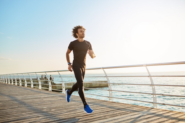 Atleta do sexo masculino atraente vestindo roupas esportivas pretas elegantes e tênis azuis. Figura de atleta homem fazendo exercícios aeróbicos em uma manhã ensolarada de verão.