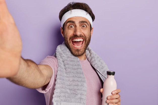 Atleta feliz estende a mão e tira uma selfie durante o treino, segura a garrafa de água, se mantém hidratado e saudável, usa uma toalha no pescoço isolada na parede roxa