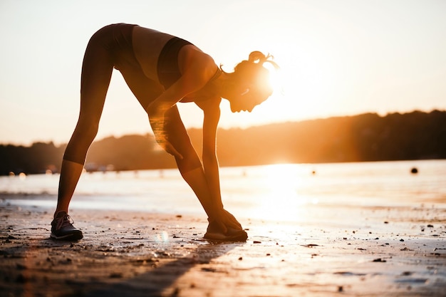 Atleta feminina fazendo exercícios de relaxamento enquanto se exercita na beira do rio ao pôr do sol