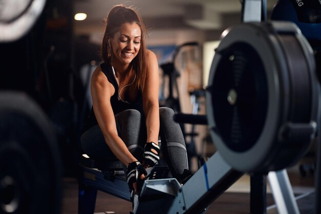 Atleta feminina feliz ajustando a máquina de remo e ouvindo música em fones de ouvido durante o treinamento esportivo em uma academia