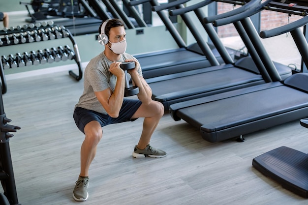 Atleta masculino com máscara facial usando o peso da mão ao fazer agachamentos no health club