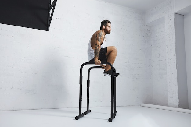 Atleta masculino com tatuagem forte em camiseta branca sem etiqueta sem rótulo mostra movimentos calistênicos Chute para fora L Sente-se, mova-se ou enfie-se em barras paralelas