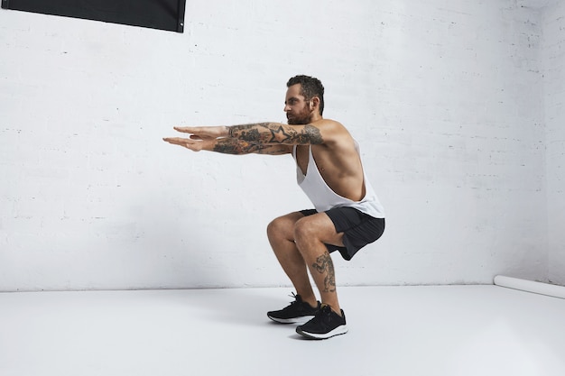 Atleta masculino com tatuagem forte em camiseta branca sem rótulo mostra movimentos calistênicos exercício de agachamento na prancha