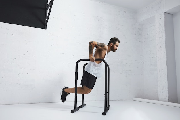 Foto grátis atleta masculino forte tatuado em camiseta branca sem rótulo sem rótulo mostra movimentos calistênicos segurando a posição de mergulho nas barras paralelas