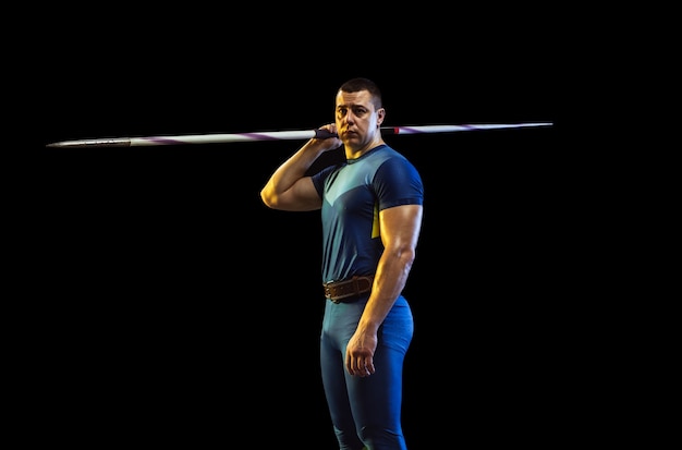 Atleta masculino praticando no lançamento de dardo em fundo preto em luz de néon. desportista profissional posando confiante. conceito de estilo de vida saudável, movimento, atividade, competição. copyspace.