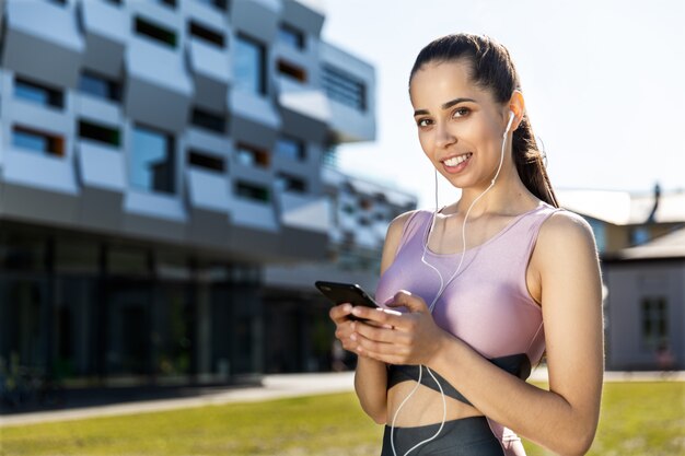 Atlética jovem está sorrindo e olhando em linha reta, com um telefone celular nas mãos e fones de ouvido, perto de edifício moderno