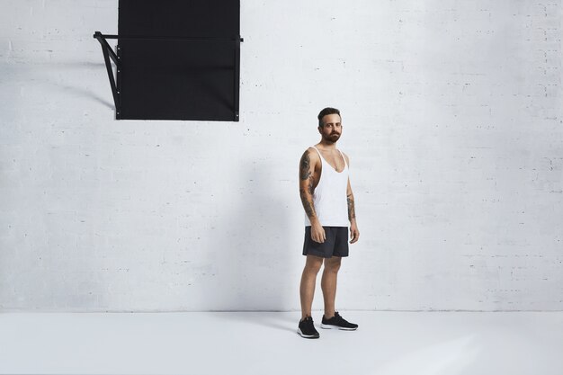 Atlético brutal e tatuado jovem em t-shirt do tanque em branco liso em pé perto da barra de pull-up em frente a parede de tijolos de grunge no ginásio branco.