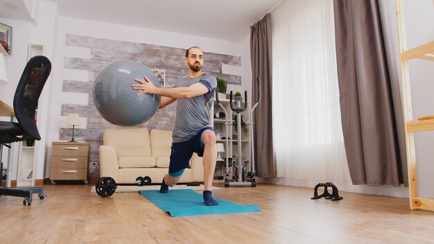 Atlético cara treinando pernas usando bola suíça em casa no tapete de ioga na sala de estar.