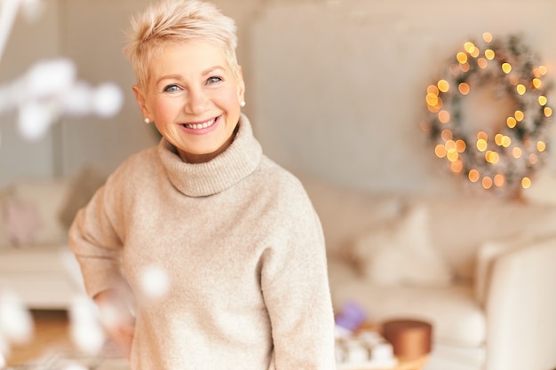 Atmosfera festiva, conceito de férias de dezembro e natal. mulher madura feliz, de cabelos curtos, confiante em um pulôver elegante, fazendo os preparativos para o ano novo, decorando a sala de estar e sorrindo com alegria