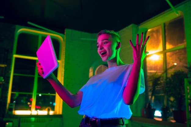 Atônito. Retrato cinematográfico de mulher elegante no interior iluminado por néon. Tons de efeitos de cinema, cores neon brilhantes. Modelo caucasiano usando tablet em luzes coloridas dentro de casa. Cultura jovem.