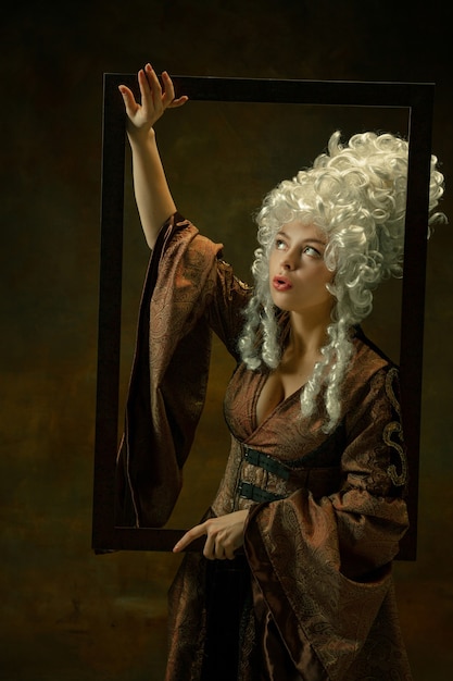 Atônito. Retrato de uma jovem medieval em roupas vintage, com moldura de madeira em fundo escuro. Modelo feminino como duquesa, pessoa real. Conceito de comparação de eras, moderno, moda, beleza.