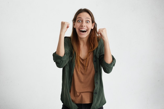 Atordoar a mulher bonita em roupas casuais, comemorando sua vitória, levantando as mãos cerrando os punhos tendo felicidade e satisfação.