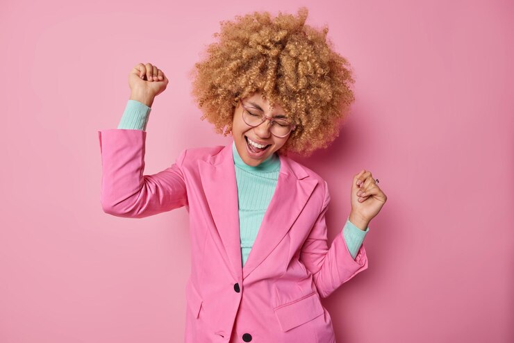Foto grátis atraente alegre mulher de cabelo encaracolado faz a dança do triunfo parecer vencedor vestida com roupas elegantes celebra conquistas pessoais isoladas sobre fundo rosa finalmente alcancei meu objetivo