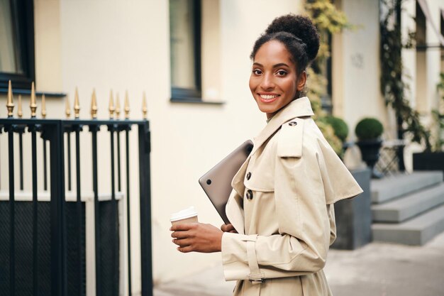 Atraente e sorridente empresária afro-americana casual em elegante casaco de trincheira com laptop e café para ir alegremente olhando para a câmera ao ar livre