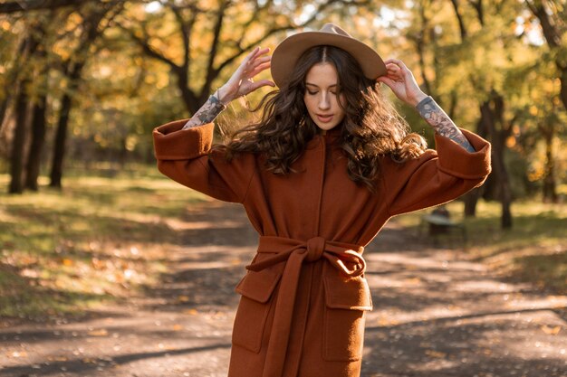 Atraente elegante e sorridente mulher magra com cabelo encaracolado caminhando no parque, vestida com um casaco marrom quente