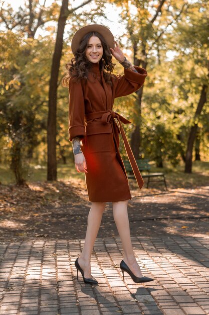 Atraente elegante e sorridente mulher magra com cabelo encaracolado caminhando no parque, vestida com um casaco marrom quente