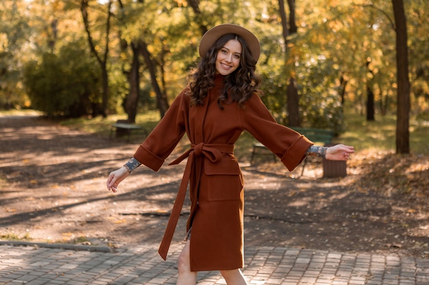 Atraente elegante e sorridente mulher magra com cabelo encaracolado caminhando no parque, vestida com um casaco marrom quente