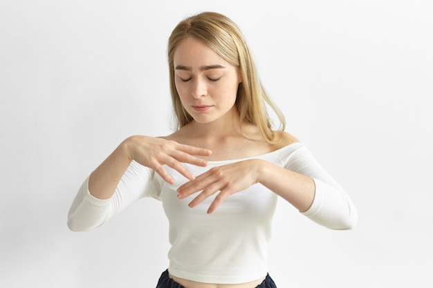 atraente elegante jovem caucasiana com top branco posando com os olhos fechados, segurando as mãos relaxadas na frente dela, movendo-se ou dançando para acalmar faixas de música relaxante