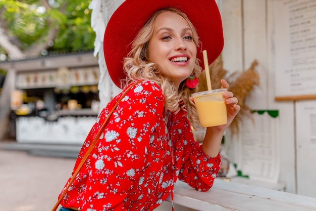 Atraente elegante loira sorridente com chapéu vermelho palha e blusa roupa da moda de verão bebendo suco de coquetel de frutas naturais