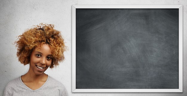 Atraente estudante universitária africana com piercing facial, apresentando seu projeto para a classe em pé na lousa em branco.