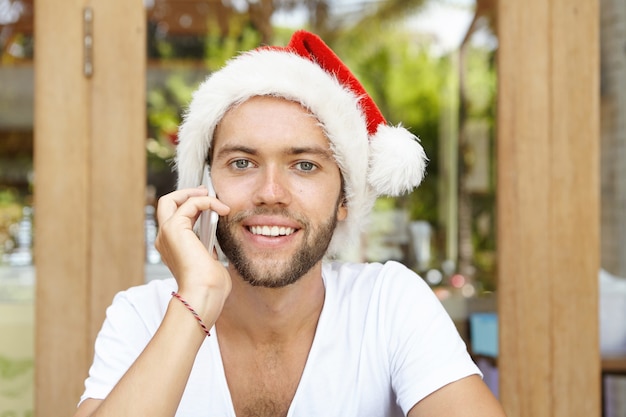 Atraente feliz jovem homem branco vestido com camiseta branca e chapéu de papai noel vermelho falando no smartphone