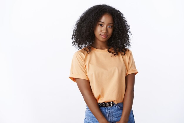 Atraente garota afro-americana estudante universitária com penteado encaracolado vestindo camiseta laranja da moda posando em uma linda parede branca, sorrindo, olhando a câmera despreocupada alegre, expressando positividade