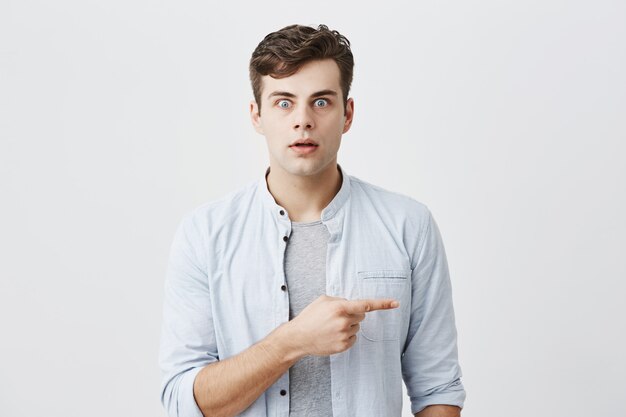 Atraente jovem homem caucasiano chocado com olhos de erros vestido casualmente apontando o dedo indicador para o lado na parede em branco cinza, mostrando algo surpreendente nele. Expressões e emoções do rosto humano